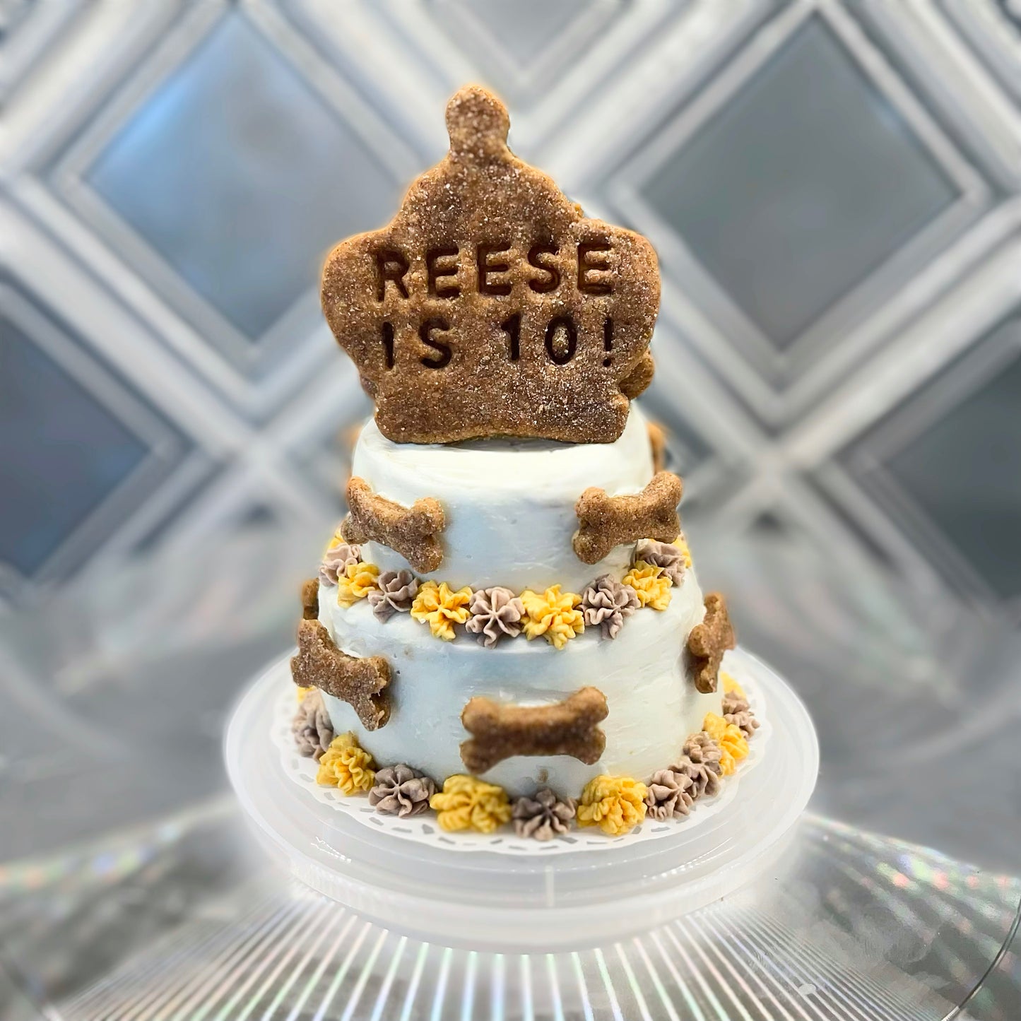 A white two-tiered cake with yellow and purple flowers and mini dog bone decorations. A cookie in the shape of a crown sits atop the cake and reads, "Reese is 10!"