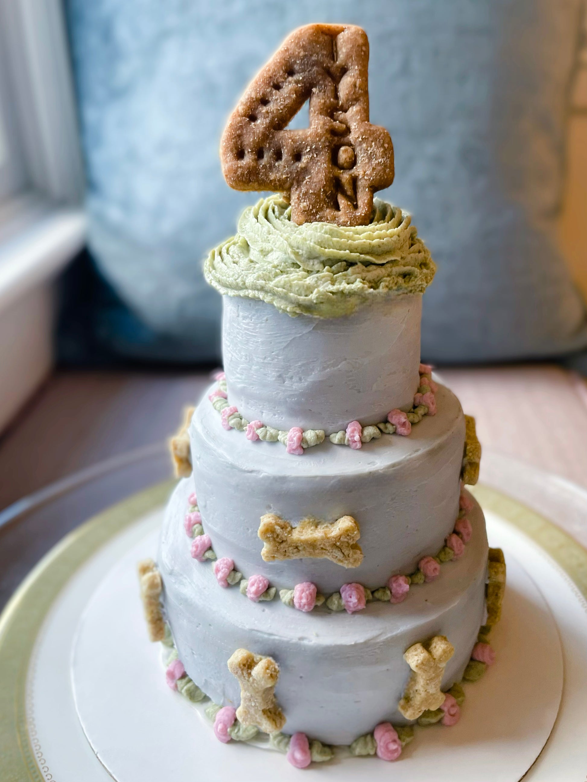 Light blue three-tier cake with light pink flowers, light green leaves, and mini dog bone cookie decorations. A cookie in the shape of the number 4 sits atop the cake, reading "Tyson.""
