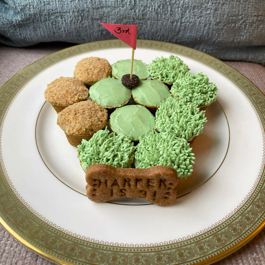 One dozen mini cupcakes in the shape of a golf course with green icing that is smooth on some cupcakes and that is spiky like grass on others. Three of the cupcakes have graham cracker crumbs on top to mimic a sand trap. There is a golf ball hole with a toothpick flag in the center and a dog cookie at the bottom of the plate that says, "HARPER IS 3!"