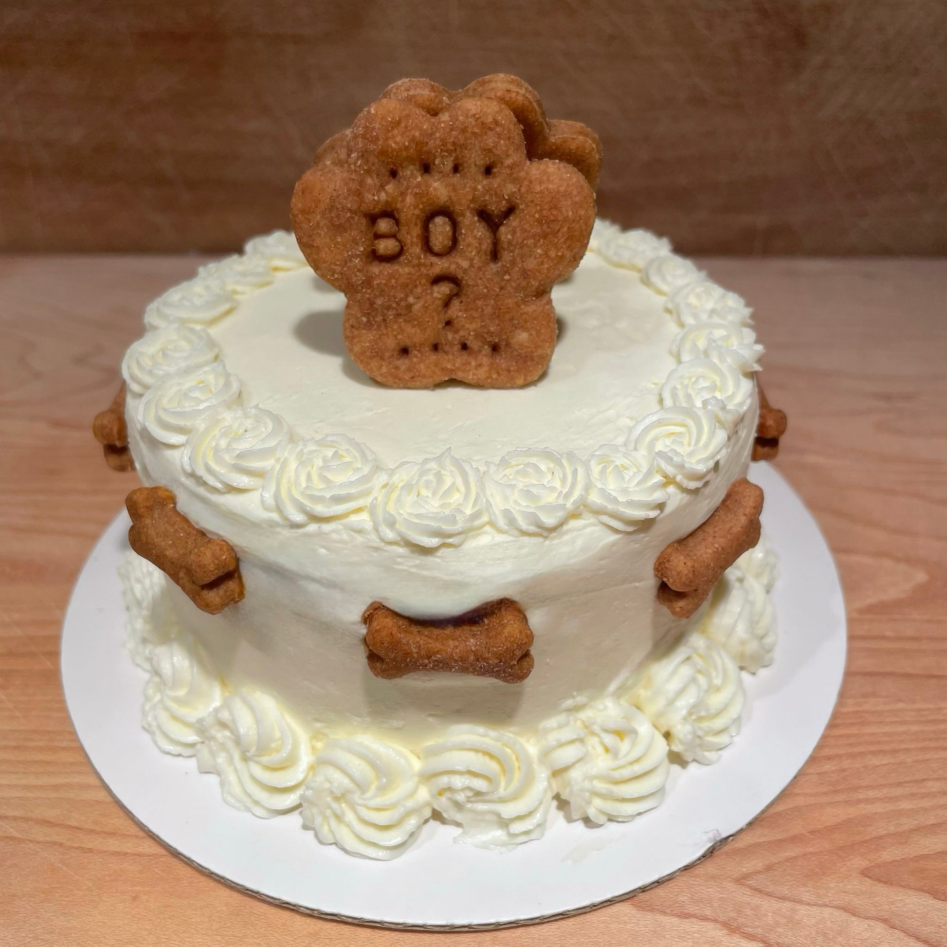 A white round gender reveal cake with white flowers on the top and bottom edges. Mini dog bone cookies surround the cake and a dog paw cookie sits atop the cake with, "BOY?," personalized onto it.