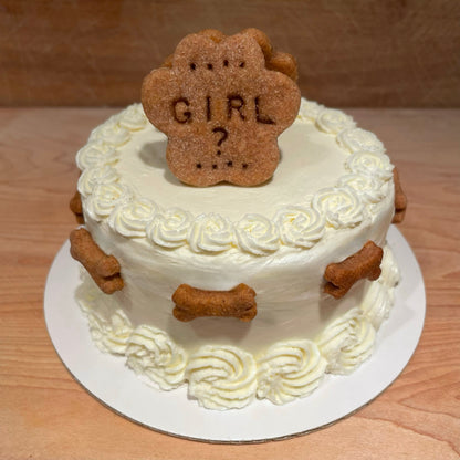 A white round gender reveal cake with white flowers on the top and bottom edges. Mini dog bone cookies surround the cake and a dog paw cookie sits atop the cake with, "GIRL?," personalized onto it.