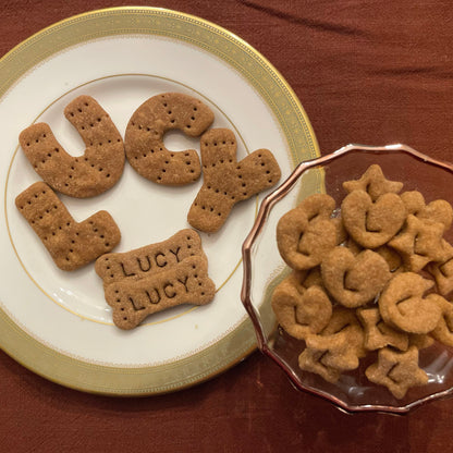 Small peanut butter dog cookies in the shapes of hearts and stars with the letter, L, stamped into each. Also, two dog bones that have been personalized with the name, "LUCY," are on the plate. Finally, each letter of the name, Lucy, has been cut into a cookie to further enhance the unique quality of this product.