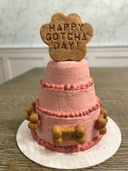 A light pink three-tier cake with slightly darker pink dots and dog bone cookie decorations. A cookie in the shape of a paw print reads, "Happy Gotcha Day!"