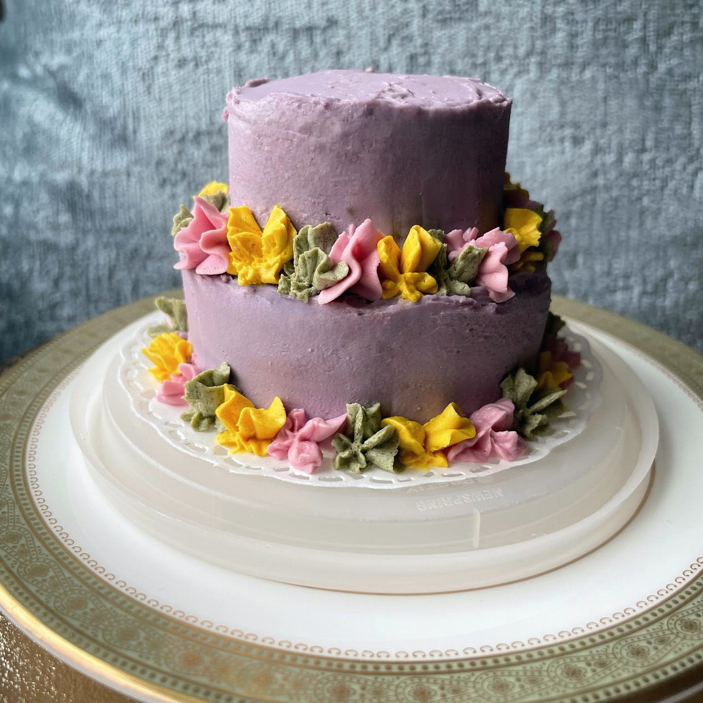 A purple two-tier cake with yellow, green, and pink flowers.