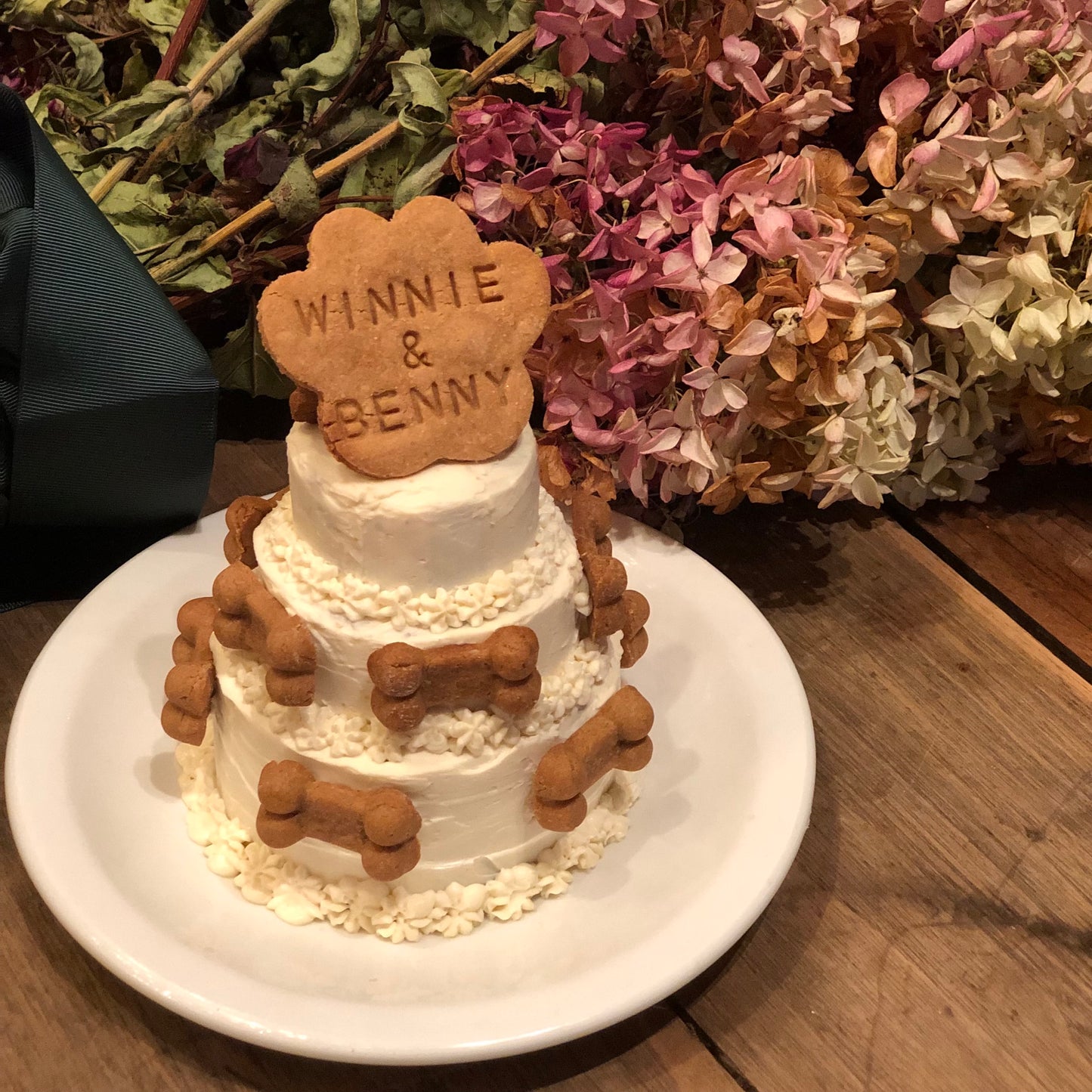 An all-white three-tier cake with white flowers and dog bone decorations. A cookie in the shape of a paw print sits atop the cake and reads, "Winnie & Benny."